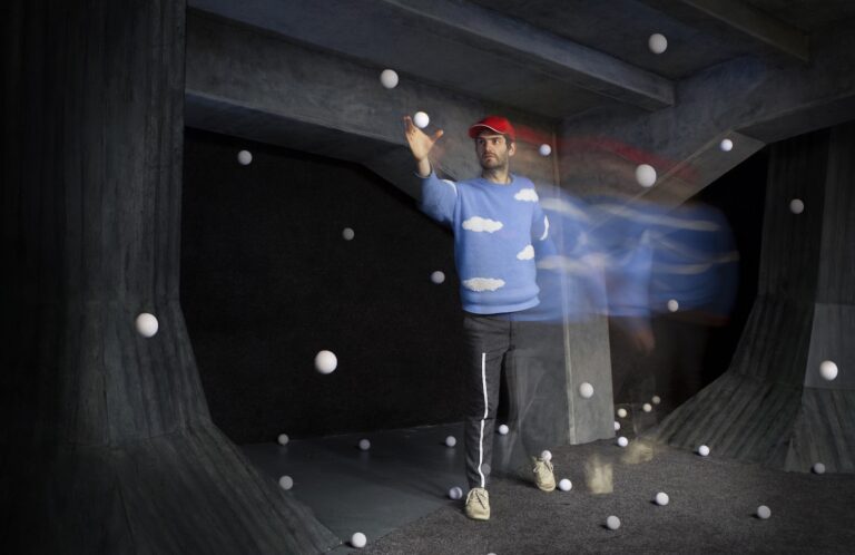 Mann mit roter Kappe und blaubem Pulli mit weißen Wolken. Um in herum fliegen viele weiße Bälle durch die Luft