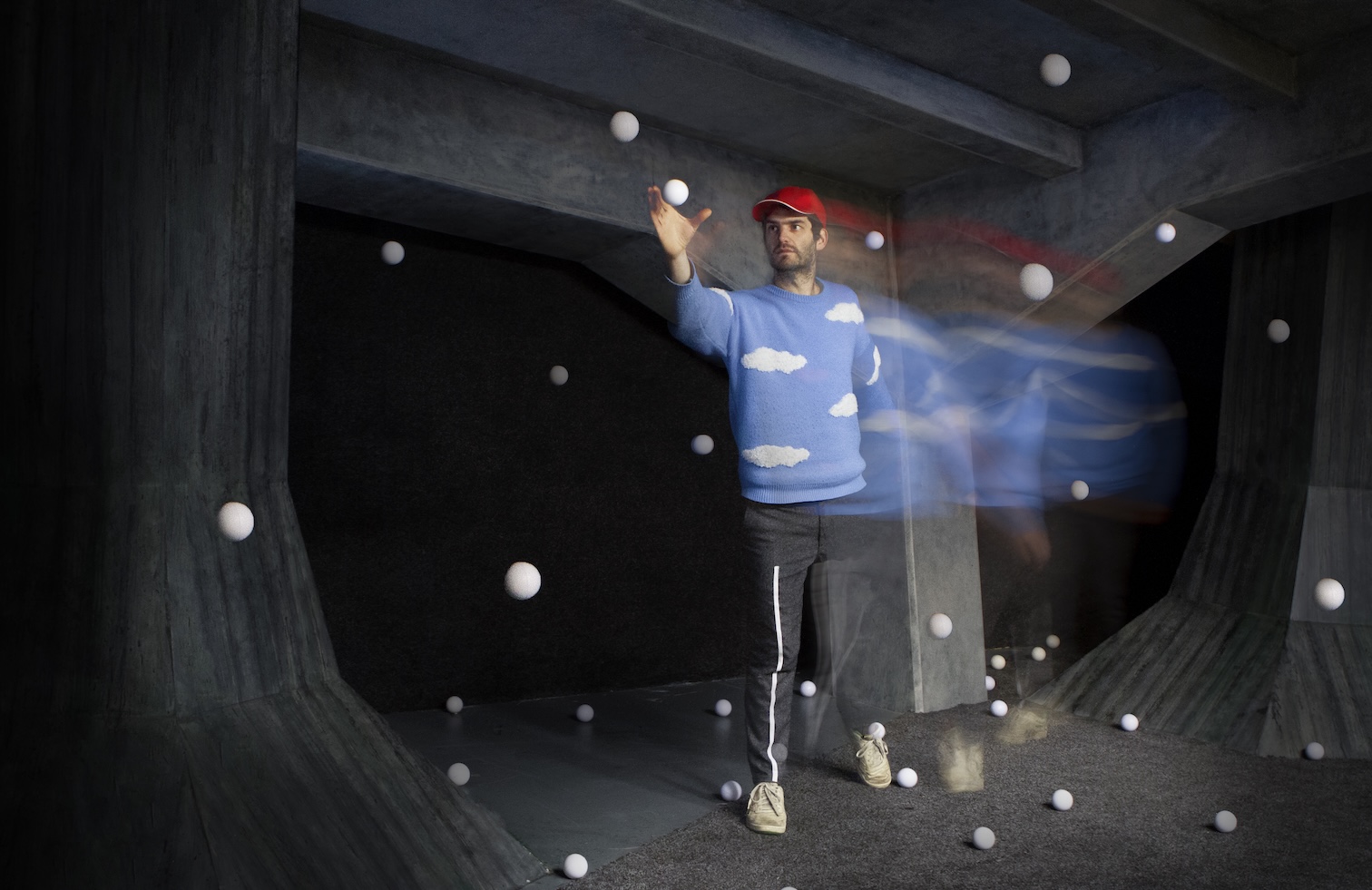 Mann mit roter Kappe und blaubem Pulli mit weißen Wolken. Um in herum fliegen viele weiße Bälle durch die Luft