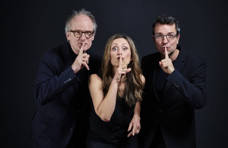 Eine Frau und zwei Männer in schwarzen Anzügen, die sich den Finger vor dem Mund legen als Zeichen zum leise sein